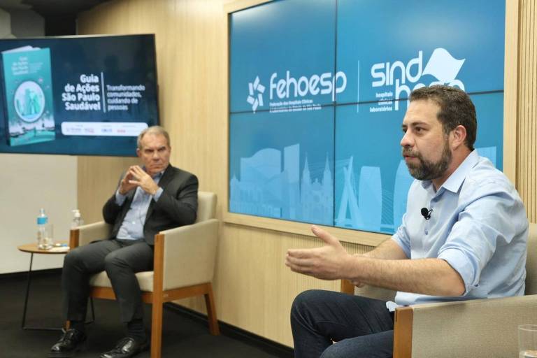 A imagem mostra um evento de discussão em um ambiente moderno. À esquerda, um homem sentado em uma cadeira, com um copo de água ao seu lado, parece estar ouvindo atentamente. À direita, outro homem, também sentado, está gesticulando enquanto fala. Ao fundo, duas telas exibem informações, uma com um gráfico e a outra com o logotipo de 'sind' e 'fehosp'. O ambiente é bem iluminado e possui paredes de madeira.