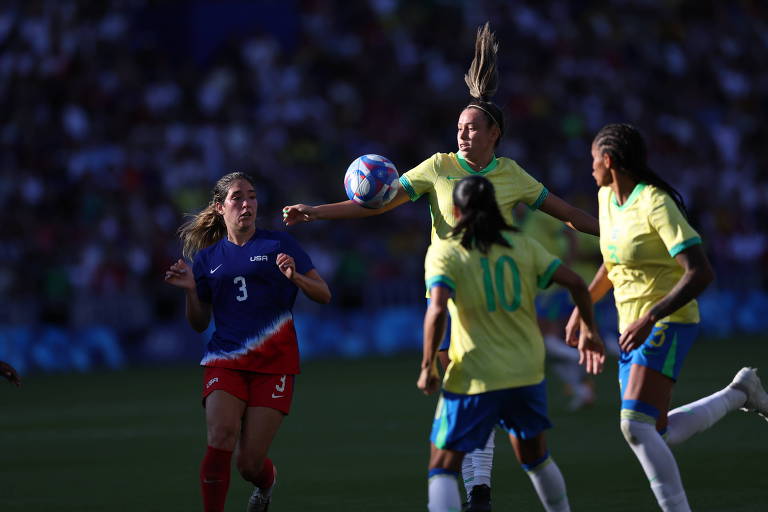 Três mulheres com camiseta verde e amarela, do Brasil, bermuda azul e meia branca estão perto de bola. uma delas está saltando. Perto, uma americana, com uniforme azum e vermelho com uma faixa branca diagonal na barriga.