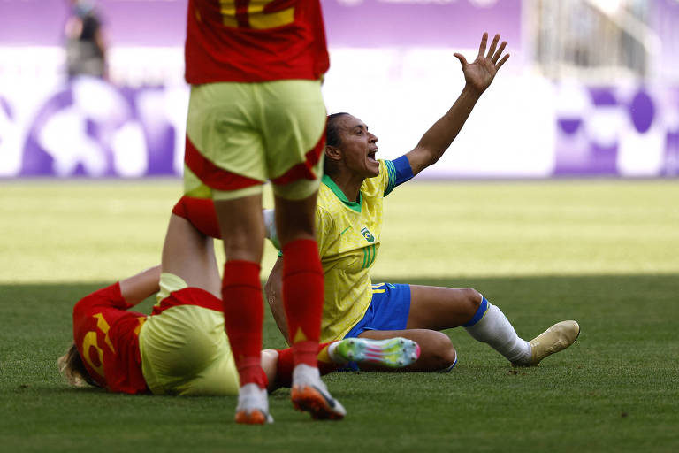 Marta, da seleção de futebol do Brasil, ergue o braço esquerdo e grita, sentada no gramado; atrás dela, jogadora da Espanha caída depois de sofrer falta da brasileira