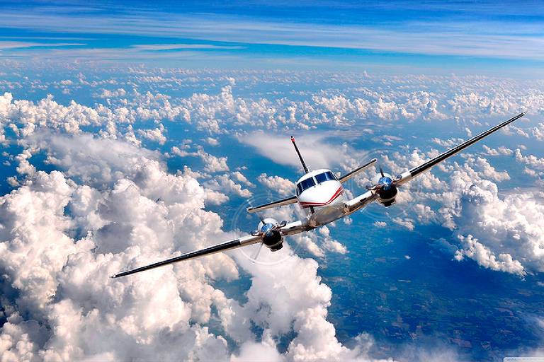 A imagem mostra um avião voando sobre um mar de nuvens brancas, com um céu azul claro ao fundo. O avião está em um ângulo que destaca suas asas e fuselagem, enquanto as nuvens parecem volumosas e fofas, refletindo a luz do sol.