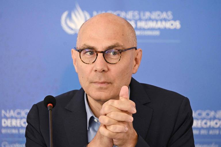 Imagem de um homem com cabelo calvo e óculos, sentado em uma mesa. Ele está olhando para frente com as mãos unidas em frente ao rosto. Ao fundo, há um painel azul com o texto 'NACIONES UNIDAS DERECHOS HUMANOS'.