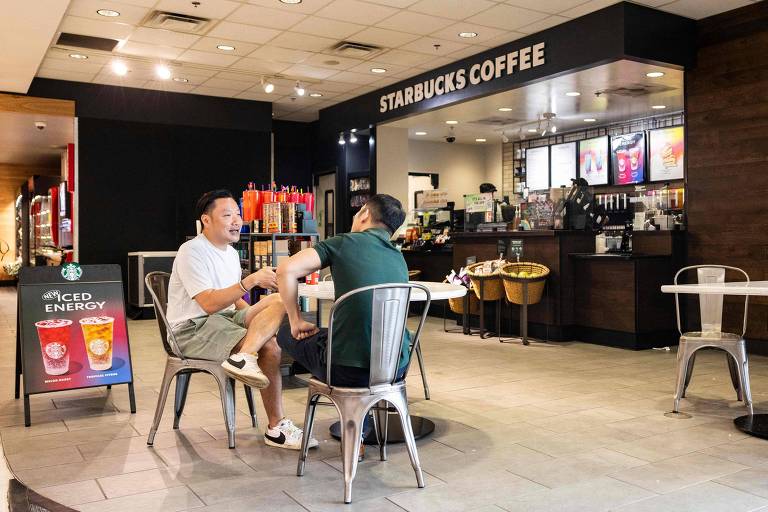 A imagem mostra duas pessoas sentadas em uma mesa em um café Starbucks. O ambiente é moderno, com mesas e cadeiras de metal. Ao fundo, há um balcão com uma variedade de bebidas e produtos expostos. O local é bem iluminado e possui uma decoração simples. As pessoas parecem estar conversando e se divertindo.