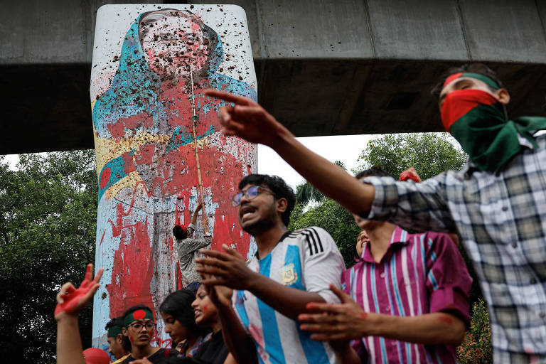 A imagem mostra um grupo de pessoas em um protesto em frente a um grande mural. O mural retrata uma figura feminina, parcialmente coberta de tinta. As pessoas estão vestindo roupas coloridas, algumas com faixas vermelhas na cabeça, e estão levantando as mãos, algumas aplaudindo. O ambiente parece ser urbano, com uma estrutura de concreto acima do mural.