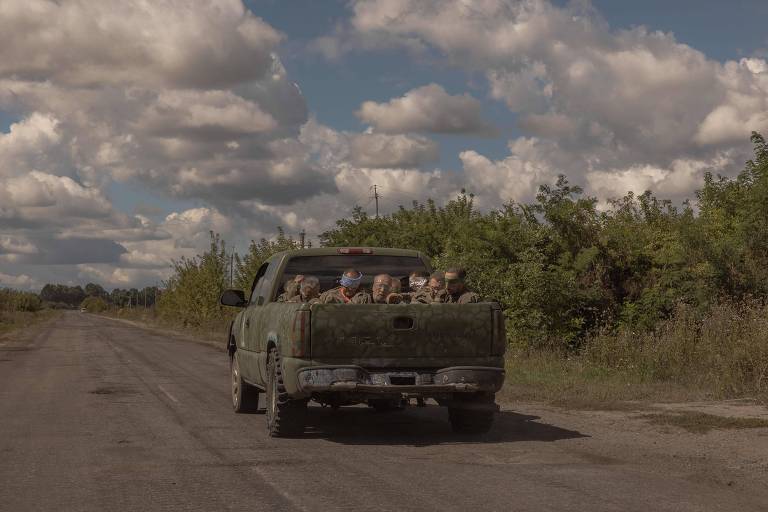 Em uma estrada, uma picape verde leva na caçamba homens com uniformes verdes com os olhos vendados por fita adesiva. O céu está cheio de nuvens, e há vegetação ao lado da pista