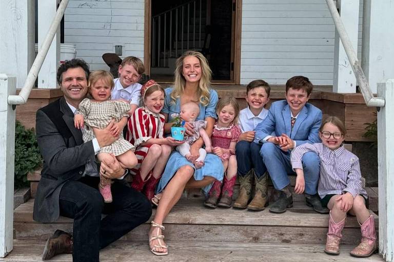 Uma família composta por um homem e uma mulher sentados em um degrau de madeira, cercados por oito crianças. O homem usa um terno escuro e segura uma criança pequena. A mulher está vestida com um vestido azul e usa sandálias. As crianças estão vestidas de maneira variada, algumas com roupas listradas e outras com vestidos. Ao fundo, há uma casa branca com uma bandeira americana pendurada. O ambiente é ao ar livre, com grama verde ao redor.