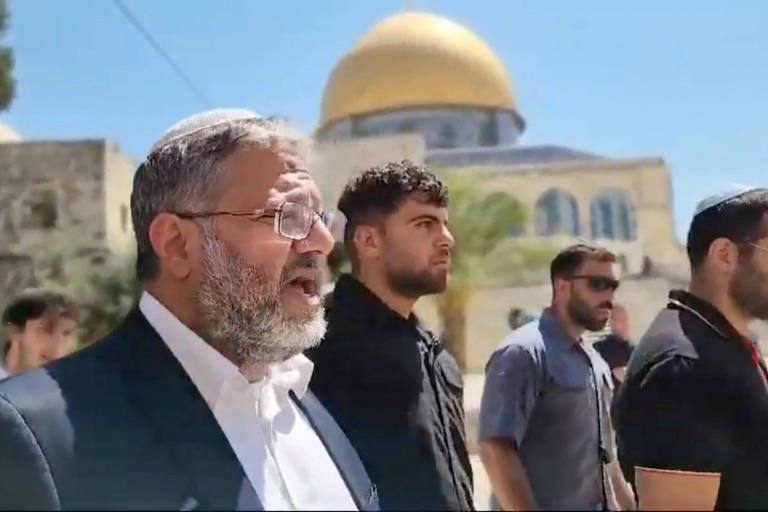 A imagem mostra um grupo de homens em pé, alguns deles falando, em frente à Cúpula da Rocha, um famoso local religioso em Jerusalém. O homem à esquerda, com barba e óculos, parece ser o mais destacado. Os outros homens estão em pé ao lado dele, com expressões sérias. O céu está claro e ensolarado, e a cúpula dourada da estrutura é visível ao fundo.