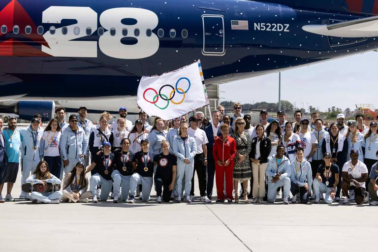 Um grupo de atletas olímpicos e oficiais posam para uma foto em frente a um avião da Delta Airlines. Eles estão vestidos com roupas esportivas, muitos usando medalhas. Um atleta segura uma bandeira com os cinco anéis olímpicos. O fundo mostra a fuselagem do avião com o número 28 e a bandeira dos Estados Unidos.
