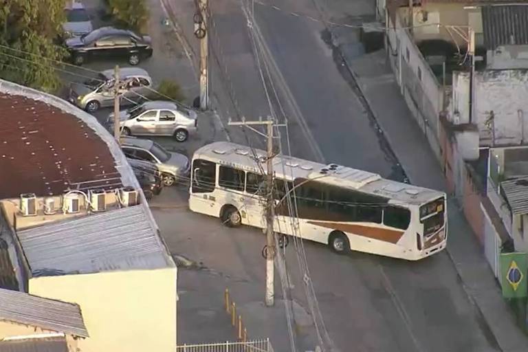 Ônibus predominantemente branco, com detalhes em marrom, parado na diagonal em uma rua vazia, sem movimentação de pessoas. Há carros estacionados por perto