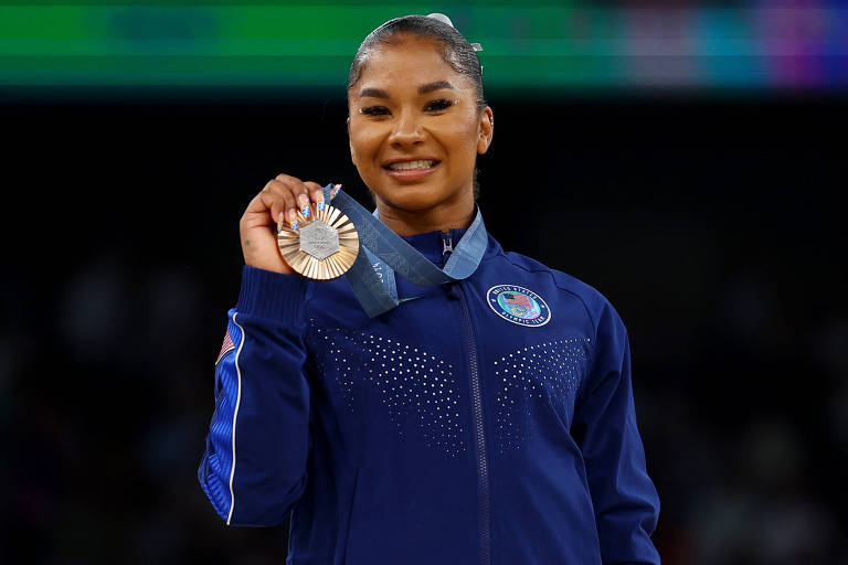 Uma atleta está sorrindo e segurando uma medalha. Ela usa uma jaqueta azul com detalhes brilhantes e um emblema que representa os Estados Unidos. A medalha é prateada com um cordão azul. O fundo é desfocado, sugerindo um ambiente de competição.
