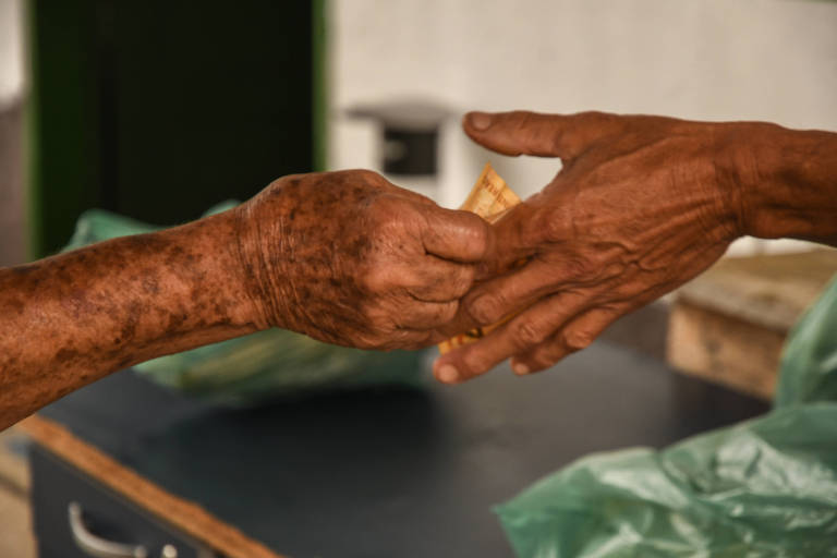 Foco em duas mãos que se tocam. uma delas é manchada, ambas são de pessoas queimadas pelo sol; uma delas, do lado esquerdo, segura uma nota de dinheiro que está parcialmente já na palma da outra mão; ao fundo se vê diversas sacolas