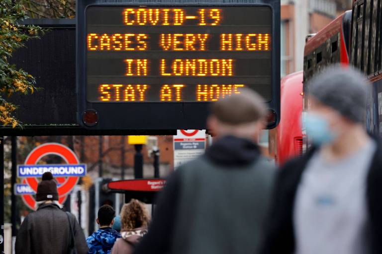 Em primeiro plano há uma placa com os dizeres "COVID-19 cases are very high in London - Stay at Home". Na frente, desfocadas, é possível ver duas pessoas, uma de costas e uma de frente usando máscara