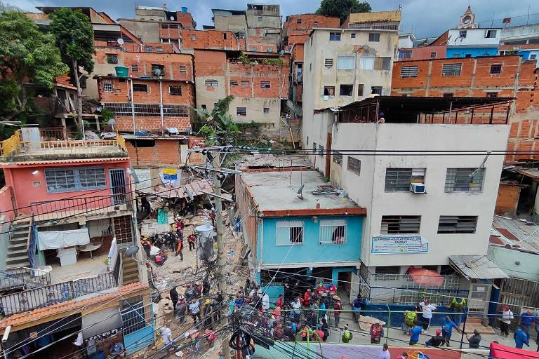 A imagem mostra uma vista aérea de uma comunidade urbana, com várias casas de tijolos expostos e estruturas de concreto. Há pessoas caminhando nas ruas, algumas em grupos, e fios elétricos visíveis cruzando a cena. O céu está nublado, e a vegetação é visível em algumas áreas. As casas são de diferentes tamanhos e cores, refletindo a diversidade da arquitetura local.