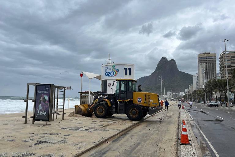 Imagem mostra máquina de limpeza de praia estacionada na areia, próxima a um ponto de ônibus. Ao fundo, há um morro característico e nuvens escuras no céu, indicando um clima nublado. A calçada ao lado da praia é visível, assim como alguns cones de sinalização