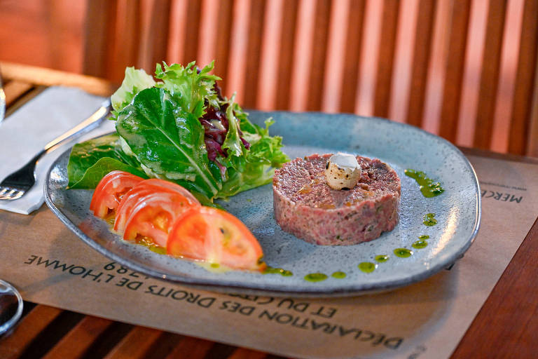 Um prato apresenta uma porção de carne moldada em forma circular, decorada com um pequeno ovo de codorna e um fio de molho verde ao redor. Ao lado, há uma salada com folhas verdes e fatias de tomate dispostas em linha. O prato é colocado sobre uma mesa de madeira, com um guardanapo e talheres visíveis ao fundo.