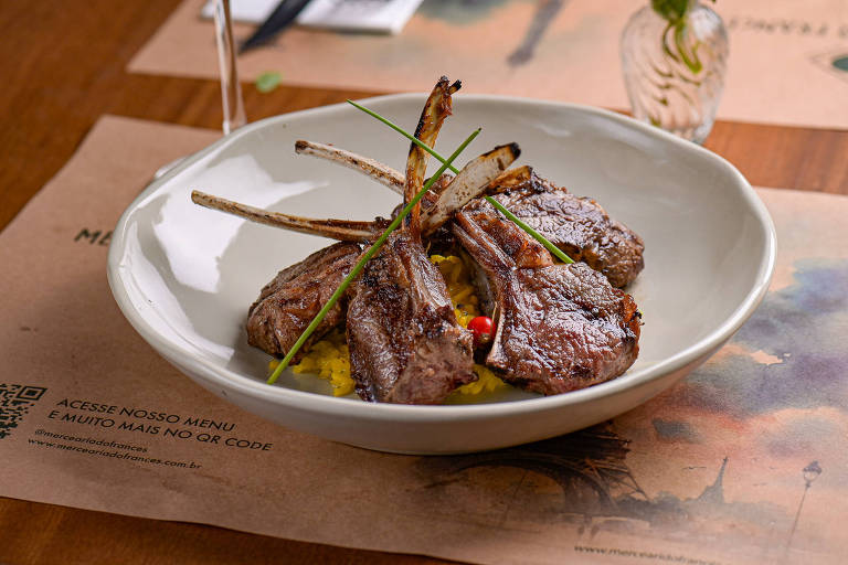 Um prato com costeletas de cordeiro grelhadas dispostas sobre uma cama de arroz amarelo. As costeletas estão decoradas com talos de cebolinha e há algumas sementes vermelhas visíveis. O fundo é uma mesa de madeira com um guardanapo e um QR code para acessar o menu.