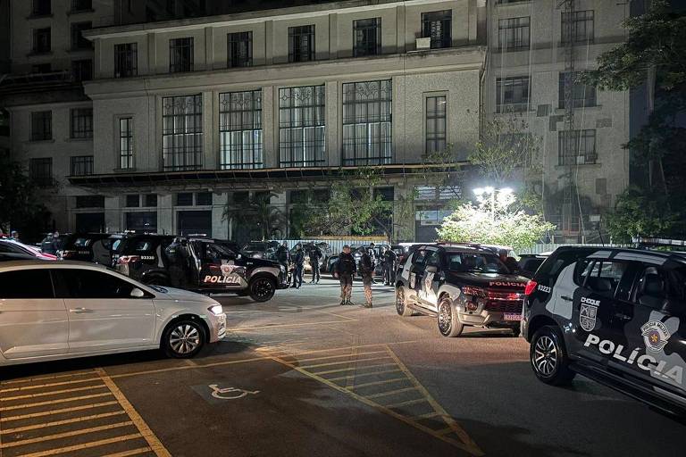 A imagem mostra uma cena noturna em um estacionamento, onde há vários veículos, incluindo carros de polícia. Um grupo de pessoas está reunido em uma área iluminada, possivelmente em uma operação policial. O ambiente é urbano, com um edifício visível ao fundo.