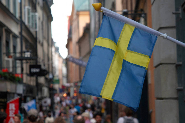 A imagem mostra uma bandeira da Suécia, com fundo azul e uma cruz amarela, pendurada em um mastro. Ao fundo, uma rua estreita e movimentada, com várias pessoas caminhando. Edifícios antigos e lojas estão visíveis nas laterais da rua, e há outras bandeiras e sinalizações ao longo do caminho.
