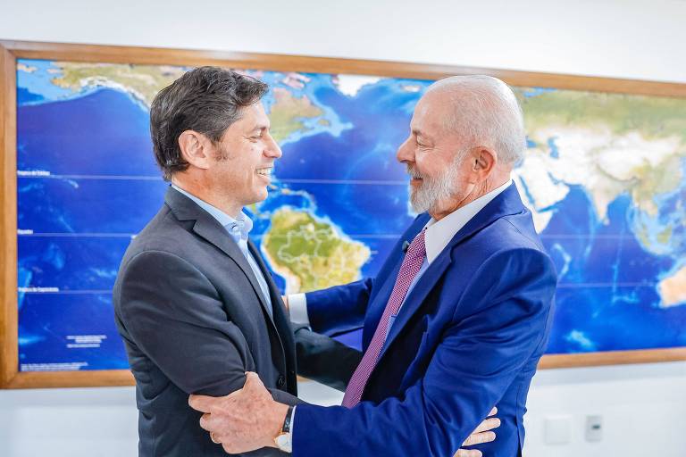 A imagem mostra Lula cumprimentando o governador de Buenos Aires, Axel Kicillof, em um ambiente interno. Eles estão de pé, um em frente ao outro, com expressões amigáveis. O homem à esquerda usa um terno escuro e o homem à direita está vestido com um terno azul e uma gravata rosa. Ao fundo, há um mapa-múndi na parede, destacando os continentes e oceanos.