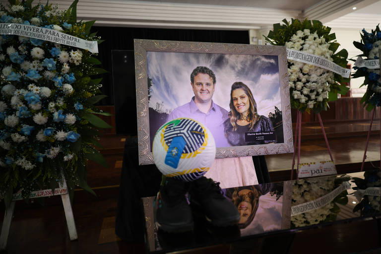 A imagem mostra uma mesa com uma foto emoldurada do casal Antônio e Kharine, rodeada por arranjos de flores brancas e azuis. Na frente da foto, há uma bola de futsal com as cores da bandeira do Brasil e um par de sapatos pretos. O ambiente é de um velório.