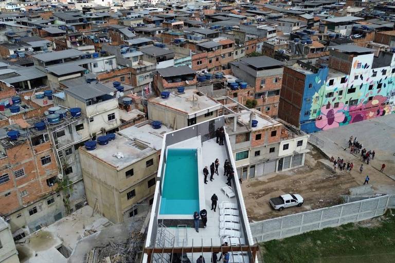 Cobertura de prédio com piscina e homens na cobertura. Ao redor do prédio, casas de dois a três andares com cor de tijolo, sem pintura na fachada