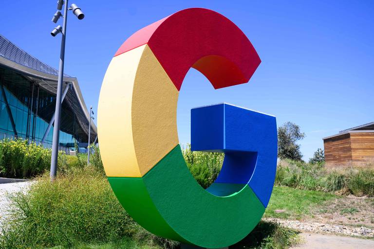 A imagem mostra uma grande escultura do logotipo do Google, composta por letras em cores vibrantes: vermelho, amarelo, verde e azul. A escultura está posicionada em um espaço externo, com um edifício moderno ao fundo e vegetação ao redor. O céu está claro e azul.
