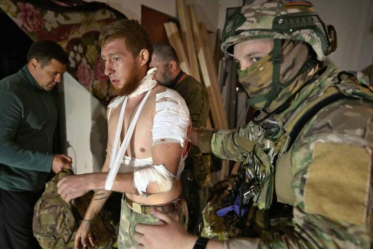 Soldado com roupa camuflada verde, capacete e rosto coberto ajuda outro homem ferido, com bandagens no braço, que está apoiado em uma faixa. Ao fundo, outro homem