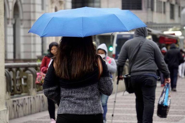 A imagem mostra uma mulher vista de costas, segurando um guarda-chuva azul. Ela está em uma rua movimentada, onde outras pessoas também caminham. O ambiente parece urbano, com prédios ao fundo e um semáforo verde visível. A mulher usa um suéter escuro e o clima parece nublado