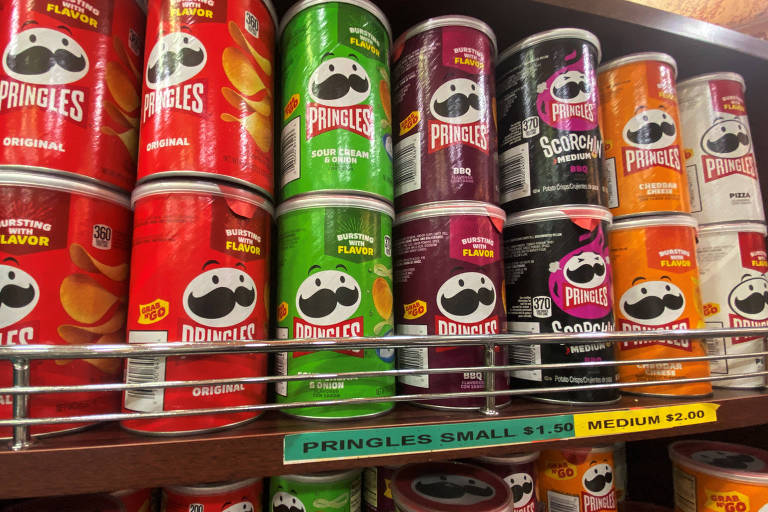 A imagem mostra uma prateleira com latas de batatas fritas Pringles. As latas estão dispostas em várias cores, incluindo vermelho, verde, roxo, preto e laranja. Cada lata possui o logotipo da marca, que apresenta um personagem com um bigode. A prateleira é de madeira e as latas estão organizadas em fileiras.