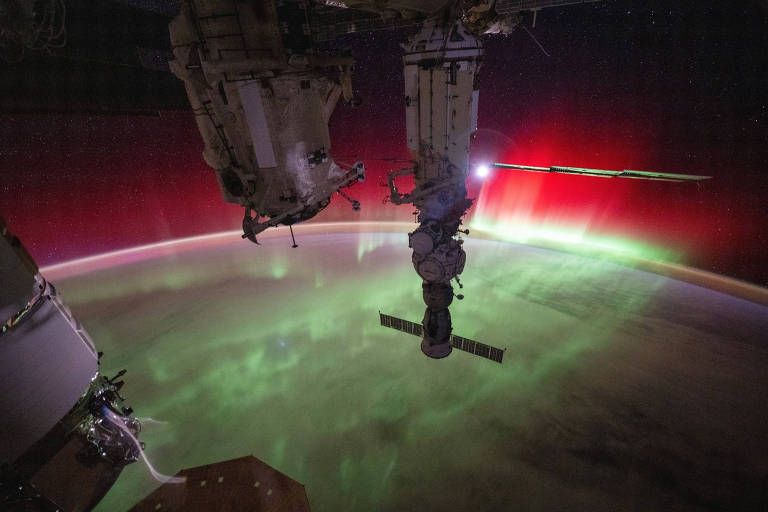 A imagem mostra uma vista do espaço com a Estação Espacial Internacional (ISS) em primeiro plano. No fundo, há uma aurora boreal com cores vibrantes, incluindo verde e vermelho, iluminando a atmosfera da Terra. As estruturas da ISS estão visíveis, com um dos módulos e painéis solares destacados contra o céu estrelado.
