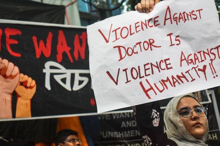 Uma mulher segurando um cartaz que diz 'Violence Against Doctor Is Violence Against Humanity!!' em um protesto. Ao fundo, há um banner com a frase 'WE WANT' e punhos cerrados, simbolizando resistência. A mulher usa um hijab e óculos, e parece estar em um ambiente urbano.