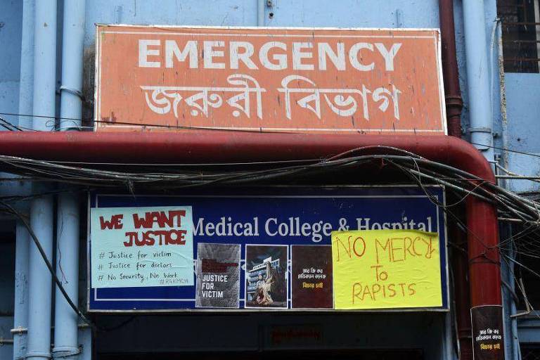 A imagem mostra a entrada de um hospital com uma placa grande em cima que diz 'EMERGENCY' em inglês e em bengali. Abaixo, há uma placa que indica 'Medical College & Hospital'. Também estão afixados cartazes com mensagens de protesto, incluindo 'WE WANT JUSTICE' e 'NO MERCY TO RAPISTS'. Fios elétricos estão visíveis ao redor da entrada.