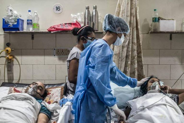 A imagem mostra um ambiente hospitalar com pacientes em camas. Dois pacientes estão deitados, usando máscaras de oxigênio. Um profissional de saúde, vestido com um traje de proteção e máscara, está atendendo um dos pacientes. Ao fundo, uma mulher está sentada, observando. O ambiente é simples, com paredes brancas e alguns equipamentos médicos visíveis.