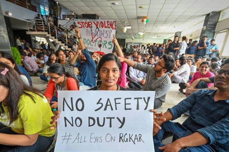 A imagem mostra um grupo de pessoas sentadas em um ambiente interno, possivelmente um hospital ou uma instituição de saúde. No primeiro plano, uma mulher segura um cartaz que diz 'NO SAFETY NO DUTY #ALL EYES ON RGKAR'. Ao fundo, há outras pessoas, algumas segurando cartazes, e uma faixa que diz 'STOP VIOLENCE AGAINST DOCTORS'. O ambiente é bem iluminado e há bandeiras tricolores decorando o espaço.