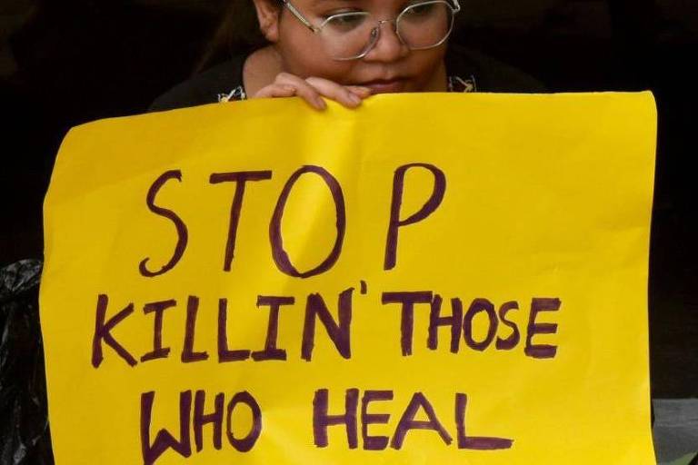 Uma pessoa segurando um cartaz amarelo com a mensagem 'STOP KILLIN' THOSE WHO HEAL' em letras roxas. A pessoa está com óculos e parece estar em um ambiente de protesto.