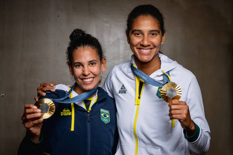 Duas atletas brasileiras posam para a foto, cada uma segurando uma medalha. A atleta à esquerda usa um agasalho azul escuro com detalhes em amarelo e exibe uma medalha dourada. A atleta à direita veste um agasalho branco com detalhes em verde e amarelo, segurando uma medalha prateada. Ambas sorriem e estão em um ambiente com parede de concreto ao fundo.