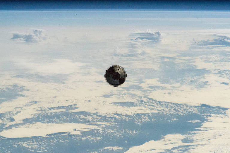 A imagem mostra a cápsula flutuando no espaço, com a Terra ao fundo. A superfície terrestre é visível com nuvens brancas espalhadas e áreas de oceano azul. O horizonte da Terra é curvado.