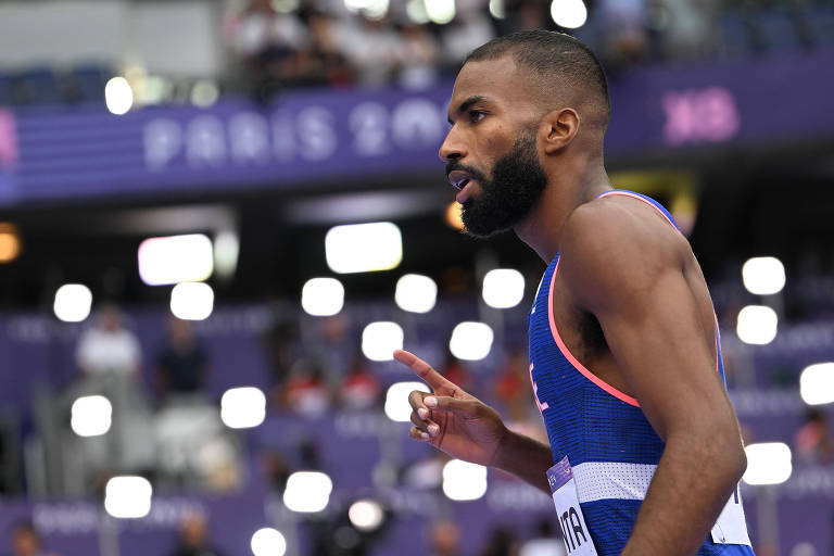 Um atleta masculino está em uma pista de atletismo, com uma expressão concentrada. Ele usa um uniforme azul com detalhes em rosa e tem uma barba bem aparada. O fundo mostra uma multidão e luzes brilhantes, sugerindo um evento esportivo importante, possivelmente uma competição internacional.
