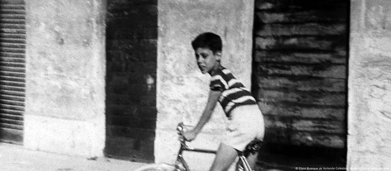 A imagem em preto e branco mostra um menino montado em uma bicicleta. Ele está usando uma camiseta listrada em preto e branco e shorts claros