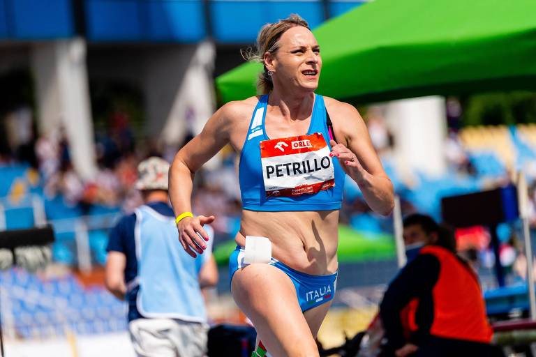 A imagem mostra uma atleta correndo em uma pista de atletismo. Ela está vestindo um uniforme azul com o nome 'PETRILLO' visível em um número de identificação. O fundo apresenta uma arquibancada com espectadores e uma tenda verde. A atleta parece concentrada e determinada durante a corrida.
