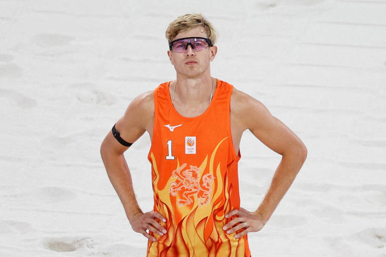 Um jogador de vôlei de praia está em pé na areia, usando uma camiseta laranja com detalhes em chamas e o número 1. Ele usa óculos escuros e tem um bracelete preto no braço esquerdo. O fundo é composto por areia clara.
