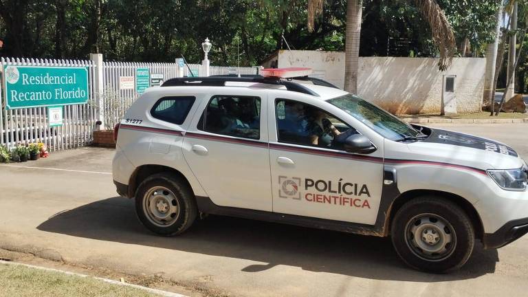 A imagem mostra um veículo da Polícia Científica estacionado em frente ao portão de um residencial chamado 'Recanto Florido'. O carro é um modelo SUV, com pintura branca e detalhes em preto e vermelho. No teto, há uma luz de emergência vermelha. Ao fundo, é possível ver o portão do residencial, que possui placas informativas e um jardim com flores