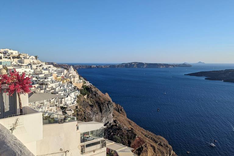 A imagem mostra uma vista panorâmica da costa de Santorini， na Grécia. À esquerda， há uma série de casas brancas empilhadas em uma encosta， com algumas flores rosas em primeiro plano. O mar azul se estende até o horizonte， onde pequenas ilhas podem ser vistas ao fundo. O céu está limpo e ensolarado， criando uma atmosfera tranquila.