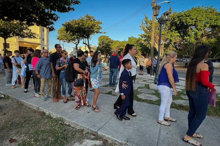 A imagem mostra uma fila de pessoas em um calçadão, com árvores ao fundo e um céu azul. As pessoas estão vestidas de maneira variada, algumas em trajes formais e outras em roupas casuais. Um menino em um terno preto está na frente da fila, enquanto os demais aguardam em pé, conversando e olhando ao redor.