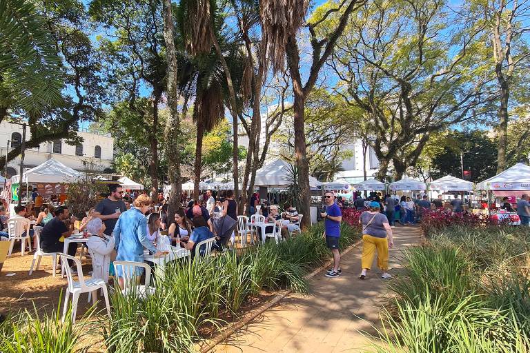 A imagem mostra uma feira ao ar livre em um parque, com várias barracas brancas dispostas ao fundo. Muitas pessoas estão sentadas em mesas brancas, algumas consumindo alimentos e bebidas. O ambiente é ensolarado, com árvores ao redor e um céu azul. Há um caminho de terra entre as barracas e a vegetação baixa na frente.