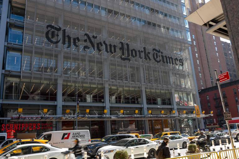 A imagem mostra a fachada do edifício do The New York Times, com o nome da empresa em letras grandes e visíveis. Na parte inferior, há uma rua movimentada com vários carros, incluindo táxis amarelos. Algumas pessoas estão caminhando na calçada em frente ao edifício. O céu está claro e o ambiente urbano é característico de Nova Iorque