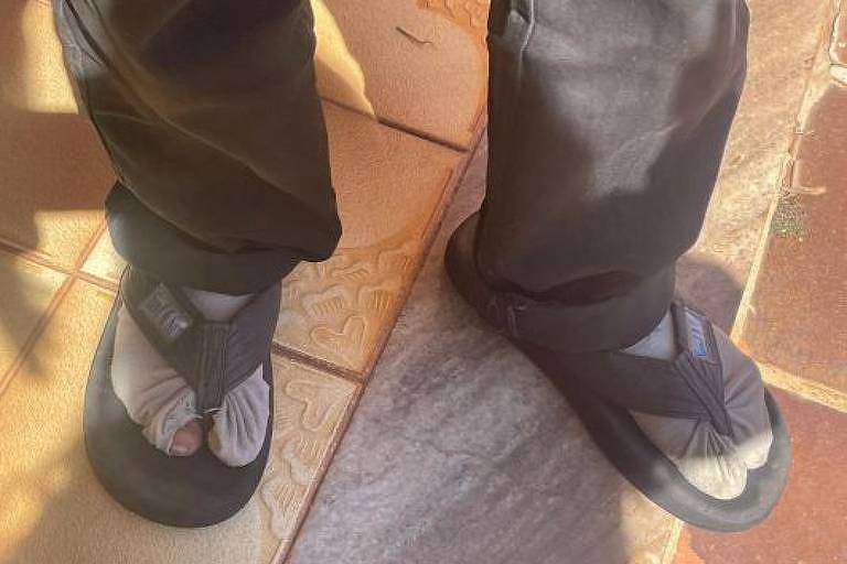 Imagem mostra dois pés com meias cinzas e chinelo preto, e parte de uma calça preta