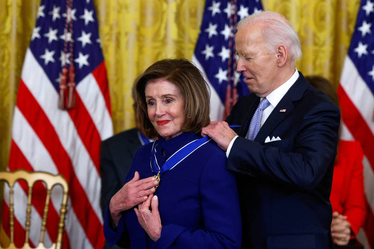 Na imagem, o presidente Joe Biden está colocando uma medalha no pescoço de uma mulher, que parece emocionada. Ao fundo, há bandeiras dos Estados Unidos e outras pessoas presentes na cerimônia. A mulher está vestindo um paletó azul e tem cabelo castanho claro. O ambiente é formal, com uma decoração elegante