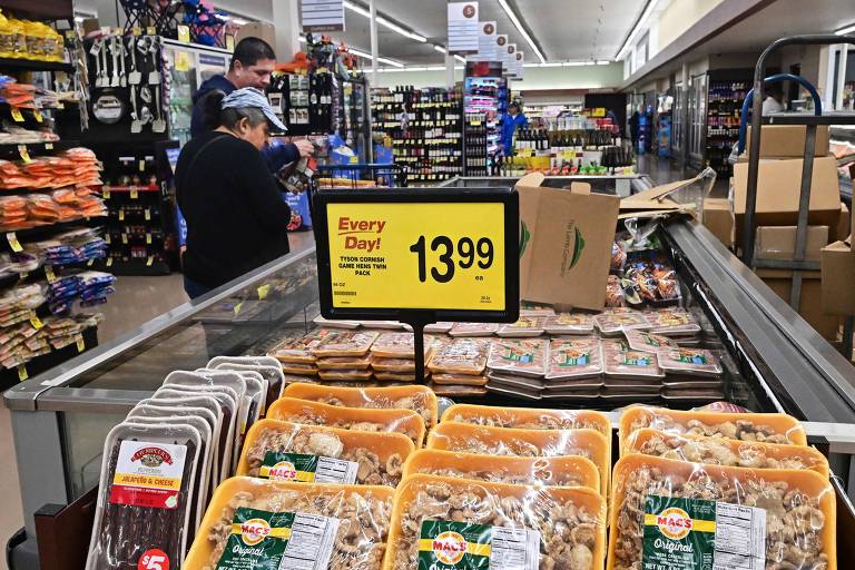 A imagem mostra o interior de um supermercado, onde duas pessoas estão fazendo compras. Em primeiro plano, há uma exibição de pacotes de carne, com um sinal amarelo destacando o preço de '13,99' para um pacote de frango. Ao fundo, prateleiras estão cheias de produtos variados, incluindo bebidas e outros itens alimentícios.
