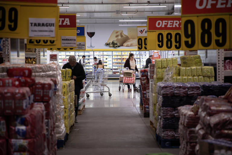 A imagem mostra o interior de um supermercado, com prateleiras cheias de produtos empilhados em sacos coloridos. Há várias pessoas fazendo compras, algumas empurrando carrinhos. Ao fundo, há uma seção de refrigerados com produtos visíveis. Placas de ofertas estão penduradas em várias partes, destacando preços. O ambiente é bem iluminado.
