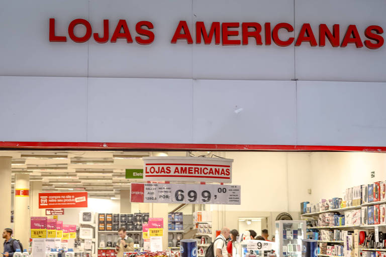 A imagem mostra a entrada de uma loja chamada 'Lojas Americanas'. Acima da entrada, está escrito 'LOJAS AMERICANAS' em letras grandes e vermelhas. Abaixo, há um cartaz com o preço de R$ 699,00. Dentro da loja, é possível ver prateleiras com produtos variados e algumas pessoas fazendo compras.